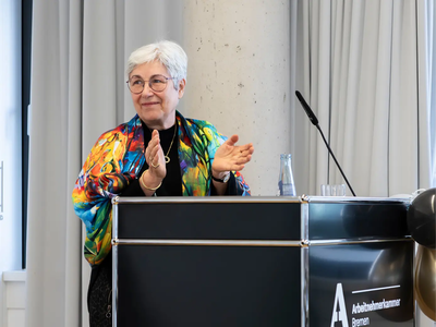 Sybille Böschen hielt die Begrüßungsrede | © Simone Hryzyk/AWO Bremerhaven Sybille Böschen steht am Redepult und klatscht in die Hände | © Simone Hryzyk/AWO Bremerhaven